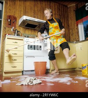 Jour de nettoyage en 1970s. Intérieur d'une cuisine où l'on voit un homme nettoyer le sol. Pour accélérer le travail, il a monté des brosses sur ses pieds. Il est acteur Stig Grybe 1928-2017. 4 octobre 1972. Kristoffersson Banque D'Images