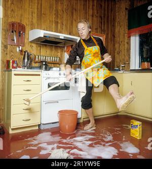 Jour de nettoyage en 1970s. Intérieur d'une cuisine où l'on voit un homme nettoyer le sol. Pour accélérer le travail, il a monté des brosses sur ses pieds. Il est acteur Stig Grybe 1928-2017. 4 octobre 1972. Kristoffersson Banque D'Images