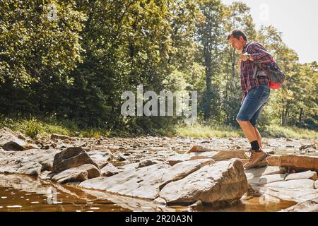 Trekking avec l'image concept de sac à dos. Backpacker portant des bottes de randonnée traversant la rivière de montagne. Homme randonnée dans les montagnes pendant le voyage d'été. Vacances Banque D'Images