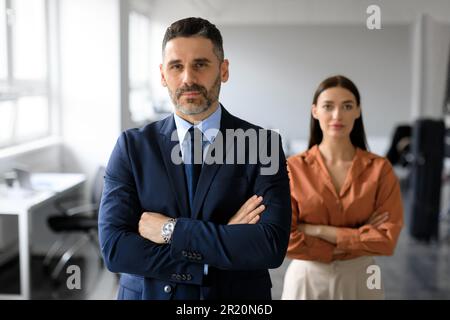 Homme d'affaires d'âge moyen et jeune femme d'affaires avec les bras croisés posant dans le bureau et regardant la caméra Banque D'Images