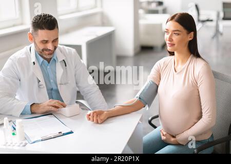 Gai gynécologue mâle mesurant la pression artérielle pour jeune femme enceinte pendant le contrôle en clinique Banque D'Images