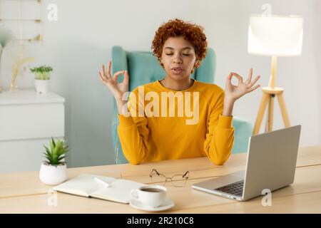 Pas de stress rester calme. Une femme d'affaires africaine consciente pratique des exercices de respiration au bureau à domicile. Une jeune femme paisible en milieu de travail aime les yeux de yoga les mains fermées dans le mouvement de mudra de menton. Méditation de bureau Banque D'Images