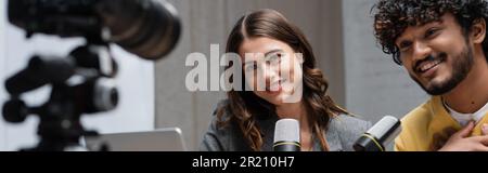 charmant télédiffuseur brunette et homme indien mauricien avec des microphones professionnels souriant près d'un appareil photo numérique flou tout en enregistrant des podcasts dans moderne Banque D'Images