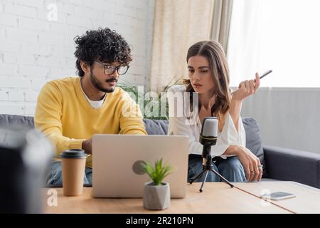 les diffuseurs interraciaux réfléchis qui regardent un ordinateur portable lors d'un appel vidéo près d'un microphone, d'un smartphone avec écran vierge, d'une boisson à emporter et d'un pot de fleurs Banque D'Images