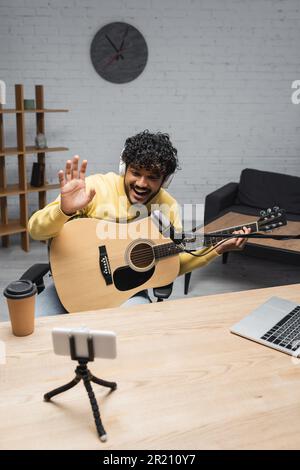 Jeune et positif, le baladoteur indien dans un casque sans fil agite la main sur un smartphone pendant un appel vidéo et tient une guitare acoustique près du café pour aller à un Banque D'Images