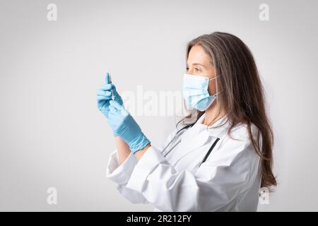 Femme senior médecin en manteau blanc, masque médical et gants de protection prenant le vaccin à la seringue, fond clair Banque D'Images