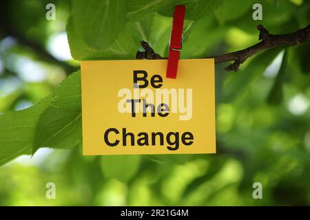 Une note de papier jaune avec l'expression être le changement sur elle attachée à une branche d'arbre avec une épingle de vêtements. Gros plan. Banque D'Images