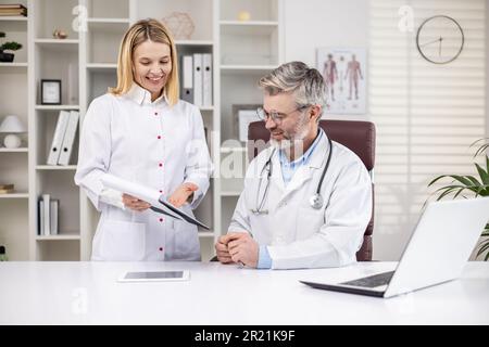 Un médecin de sexe masculin senior à cheveux gris parle avec une femme médecin, assistante. L'homme est assis au bureau, la femme montre des documents, les antécédents médicaux du patient, le diagnostic, le traitement. Banque D'Images