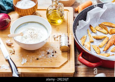 Plat à pâtisserie rempli de pommes de terre rôties ou de frites cuites dans une cuisine en désordre avec un bol de sauce au yaourt, huile d'olive, ail et épices et con Banque D'Images