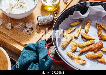 Plat à pâtisserie rempli de pommes de terre rôties ou de frites cuites dans une cuisine en désordre avec un bol de sauce au yaourt, huile d'olive, ail et épices et con Banque D'Images