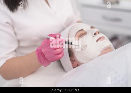 Gros plan sur les mains de esthéticiennes en gants roses application d'un masque de beauté facial à l'aide d'une brosse Banque D'Images