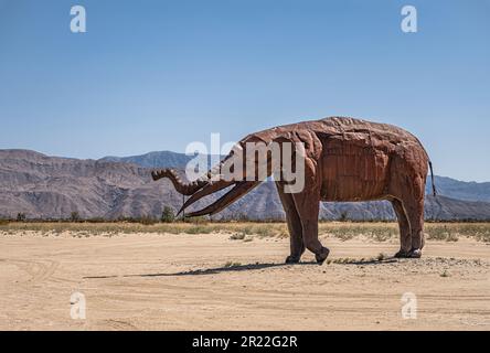 Borrego Springs, CA, Etats-Unis - 24 avril 2023 : statue de mammouth géant préhistorique en métal rouillé brun sur un sol sablonneux du désert avec des mauvaises herbes vertes sous le bleu s. Banque D'Images