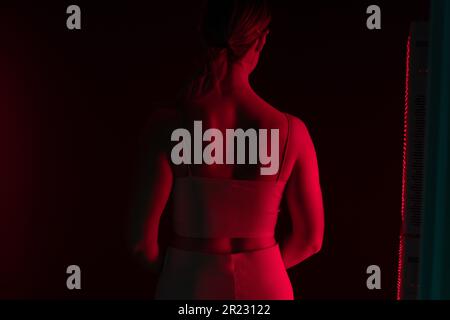 Vue arrière d'une femme recevant une luminothérapie à la lumière rouge sur son dos. Femme debout à côté d'un appareil de lumière rouge dans un spa cosmétique. Traiteurs anti-âge pour les soins de la peau Banque D'Images