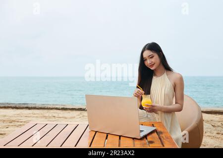 Jeune femme assise à table sur la plage, prenant un cocktail et travaillant sur un ordinateur portable Banque D'Images