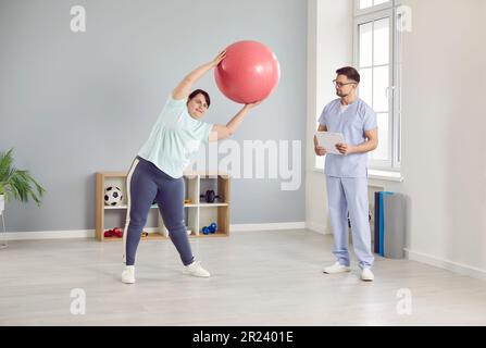 Femme en surpoids faisant des exercices avec une balle d'ajustement sous le contrôle d'un physiothérapeute Banque D'Images