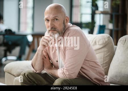 Sage homme âgé, avec une touche de gris dans la barbe, regarde directement dans l'appareil photo dans l'intérieur confortable de la maison. Sa main repose doucement sur la barbe argentée, exsurant le sens de la pensée et de la contemplation. Photo de haute qualité Banque D'Images