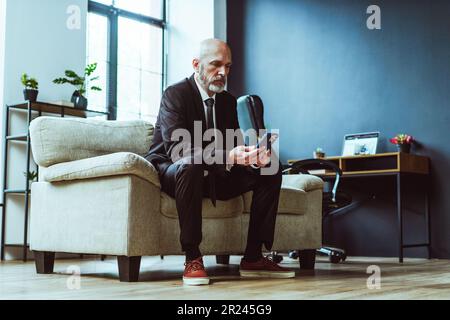 Vieux homme d'affaires vêtu d'un costume noir peut être vu soit par SMS sur le téléphone ou la navigation sur Internet. Homme mûr, avec une barbe argentée distinguée, s'assoit confortablement dans une chaise dans un bureau loft élégant. . Photo de haute qualité Banque D'Images