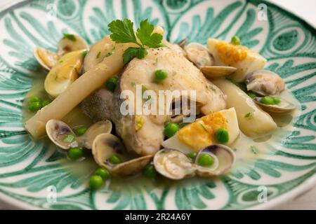 Merlu de style basque cuit avec du persil vert et de la sauce à l'ail, servi avec des asperges, des palourdes, des œufs durs et des petits pois. Banque D'Images