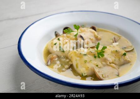 Merlu de style basque cuit avec du persil vert et de la sauce à l'ail, servi avec des asperges, des palourdes, des œufs durs et des petits pois. Banque D'Images