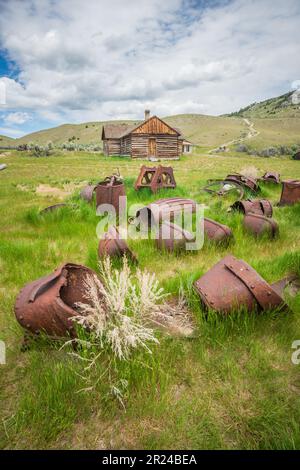 Des barils en métal rouillés et des ferrailles sont éparpillés dans l'herbe verte devant une cabane en rondins au loin sur une colline du parc d'État de Bannack, dans le Montana. Banque D'Images