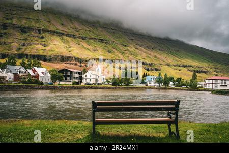 Une scène idyllique avec un banc en bois situé près d'une rivière bordée de maisons à Seydysfjordur, en Islande Banque D'Images