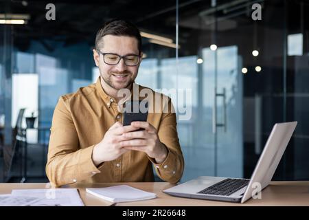 Un jeune travailleur de bureau souriant s'assoit à un bureau avec un ordinateur portable et écrit un message au téléphone, discute, vérifie le courrier, lit les nouvelles, fait une pause au travail Banque D'Images