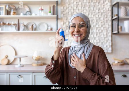 Portrait d'une jeune femme musulmane souriante dans le hijab tenant l'asthme et l'allergie inhalateur à la maison dans la cuisine, pointant et posant à la caméra. Banque D'Images