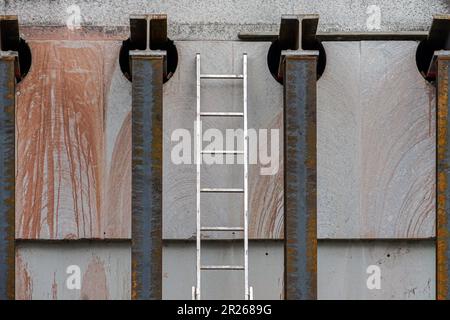 Entre deux colonnes en métal rouillé, une échelle en aluminium est penchée contre un mur de maison. Banque D'Images
