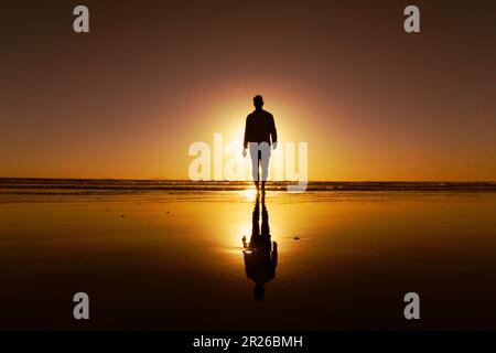 Vue à angle bas d'un jeune homme marchant sur la plage au coucher du soleil avec son ombre se reflétant dans le sable humide Banque D'Images