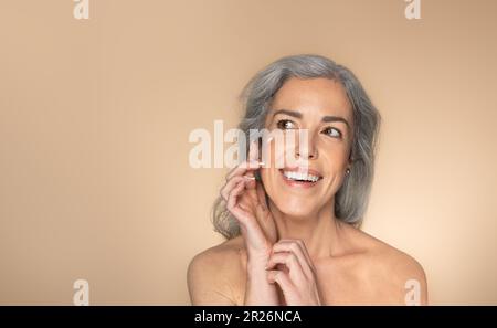 Bonne femme senior appliquant la crème du visage, prenant soin de ses regards sur fond beige studio, panorama avec espace libre Banque D'Images
