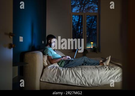 Étudiant noir allongé dans le lit avec ordinateur portable le soir, étudiant jusqu'à tard dans la journée Banque D'Images