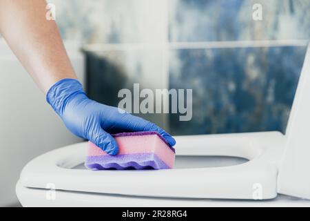 Gros plan sur la main féminine gantée de gants de protection en caoutchouc bleu. Nettoyage de la cuve cuvette de toilette, siège avec détergent liquide, lingettes humides dans la salle de bains, toilettes. Lavage des femmes, désinfection du siège des toilettes Banque D'Images