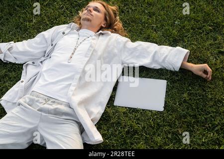 vue en grand angle du jeune homme de yoga dans des vêtements blancs couchés près d'un ordinateur portable sur un terrain vert Banque D'Images