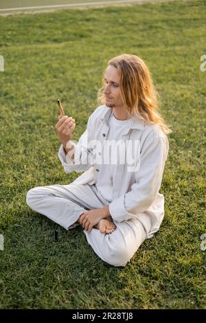 jeune homme de yoga dans des vêtements blancs regardant le bâton parfumé de palo santo tout en étant assis dans le sukhasana pose sur l'herbe verte Banque D'Images