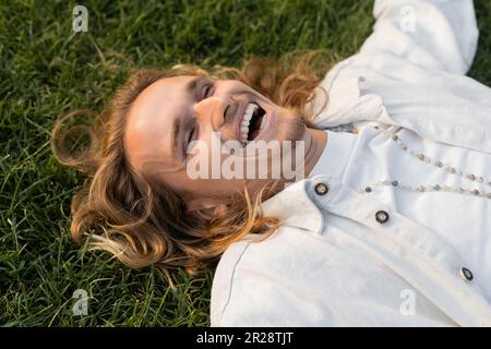 vue en grand angle d'un homme heureux en chemise blanche et perles couchée sur l'herbe verte à l'extérieur Banque D'Images