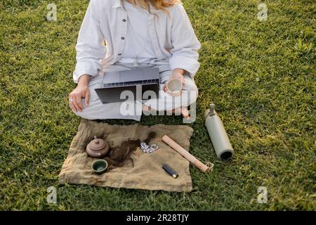 vue partielle de l'homme de yoga assis sur une pelouse verte avec un ordinateur portable et du thé plus pur près des thermos et tapis de lin avec théière en argile et perles de mala Banque D'Images