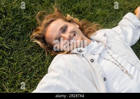 vue en grand angle d'un jeune homme gai en chemise de lin blanc, allongé sur une pelouse verte et souriant à l'appareil photo Banque D'Images