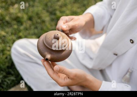 vue courte de l'homme dans des vêtements en lin blanc tenant une théière en argile tout en étant assis à l'extérieur Banque D'Images