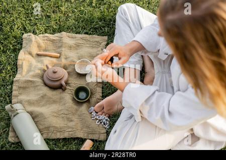 vue de dessus de l'homme de yoga tenant bâton parfumé près de tapis de lin avec théière d'argile et bols sur pelouse herbacée Banque D'Images