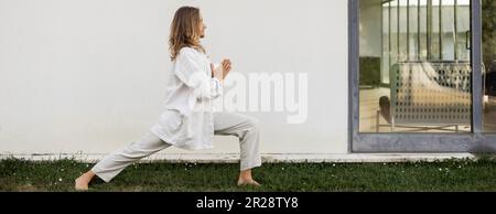 vue latérale de l'homme élégant en vêtements blancs pratiquant le yoga en posture de guerrier avec anjali mudra geste près de la maison moderne, bannière Banque D'Images