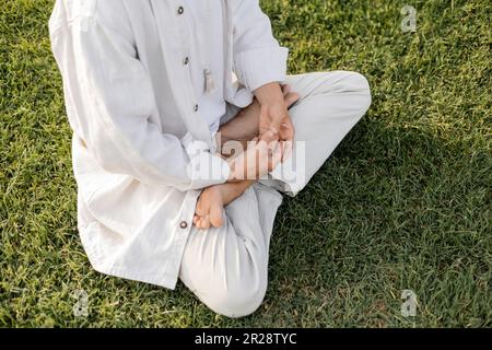 vue partielle de l'homme pieds nus dans des vêtements de lin blanc méditant dans la posture de lotus sur pelouse verte herbacée Banque D'Images