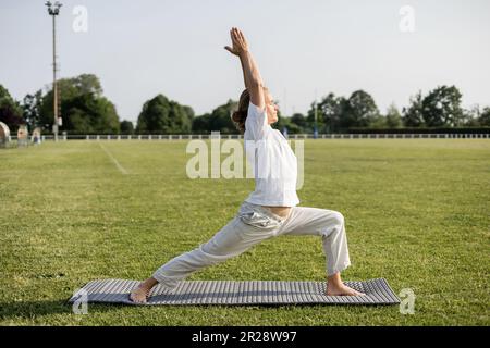 vue latérale de l'homme sportif pieds nus méditant dans la posture du guerrier avec les mains levées sur le stade à l'extérieur Banque D'Images