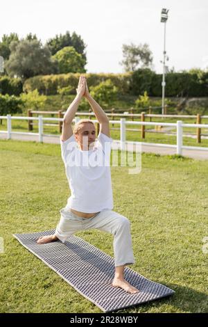 homme pieds nus dans un pantalon en lin et t-shirt blanc pratiquant le yoga dans une posture de guerrier avec des mains levées sur une pelouse herbacée Banque D'Images