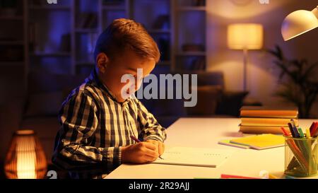 Un petit garçon brunette s'assoit à la table dans sa chambre le soir et dessine. Un pré-chooler utilise son imagination, crée une image, rêve d'être Banque D'Images