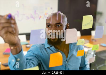 Un homme d'affaires américain africain barbu écrit des idées sur un mur de verre par des notes adhésives au bureau Banque D'Images