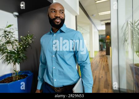 Homme d'affaires afro-américain barbu avec ordinateur portable donnant sur l'extérieur tout en marchant dans le couloir, espace de copie Banque D'Images