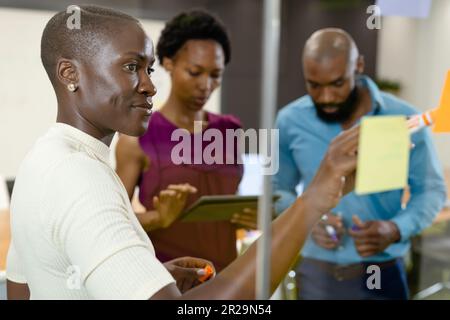 Femme afro-américaine écrivant sur une note adhésive sur un mur de verre tout en discutant avec ses collègues Banque D'Images