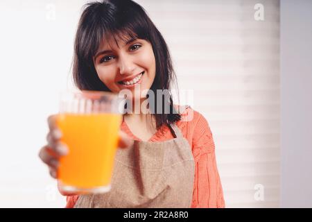 Brunette femme caucasienne avec de longs cheveux dans un tablier regarde dans la caméra, tenant un verre de jus d'orange frais, signifiant la santé et la vitamine C. Banque D'Images