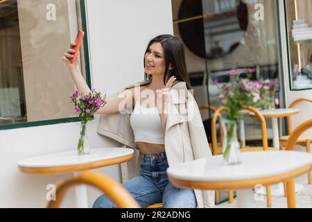 une femme heureuse avec de longs cheveux assis sur une chaise près d'une table de bistro avec des fleurs dans un vase et prenant un selfie, en train de faire des pieds-de-sac, posant dans des vêtements branchés dans un café Banque D'Images