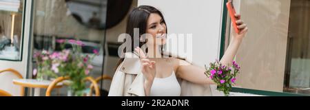 une femme heureuse avec de longs cheveux prenant selfie, en train de se mettre en porte-à-terre, assis dans des vêtements tendance avec une veste en cuir beige près de fleurs floues dans un café sur la terrasse Banque D'Images